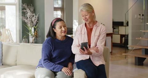 Diverse female friends chatting on sofa holding smartphone