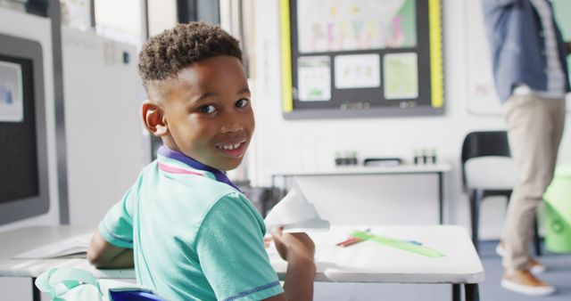Young boy with afro hair smiling and looking back in a classroom while holding paper. The bright and casual setting suggests a positive learning environment. Ideal for educational content, schools, child psychology, or back-to-school campaigns.