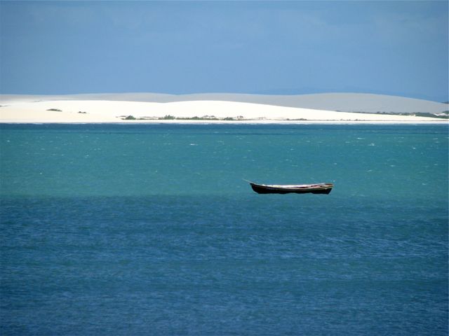 Solitary wooden boat floating on a calm blue ocean with a horizon of white sand dunes under clear blue sky. Ideal for travel blogs, nature magazines, posters about loneliness or tranquility, and backgrounds for websites promoting relaxation and vacations.