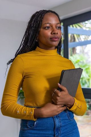 African american woman holding tablet wearing mustard turtleneck and jeans near window