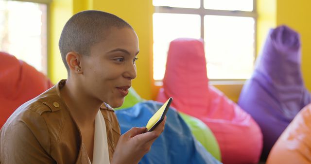 A young African American woman is using voice assistant on her phone while sitting in a room filled with colorful bean bags. Ideal for portraying modern technology usage, casual settings, communication, and relaxed work or learning environments.