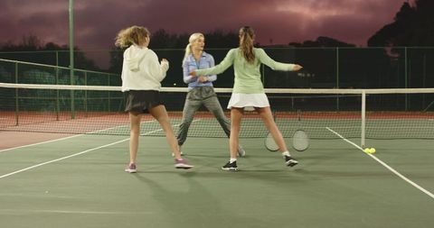 Women practicing tennis at dusk on hard court — coach leading group drill for teamwork