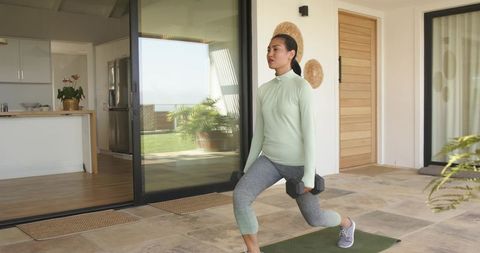 Asian woman performing dumbbell lunges on sunlit patio for at-home fitness and strength