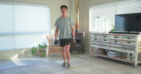 Asian man performing dumbbell lunges in sunlit minimalist living room for home fitness