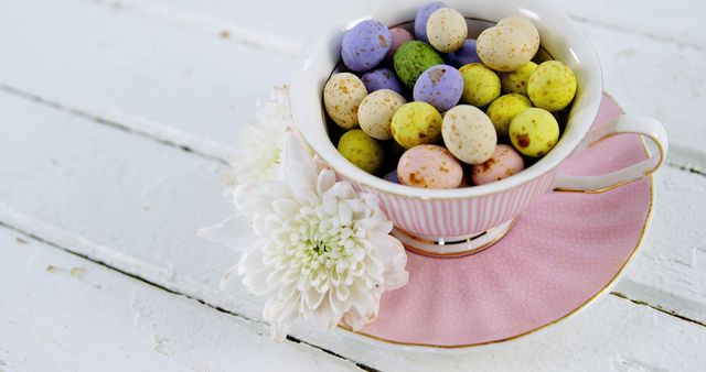 A delightful arrangement of pastel-colored chocolate eggs filling an elegant teacup with a pink saucer, set on a rustic white wooden table. White chrysanthemums add a delicate floral touch. Perfect for Easter-themed designs, holiday promotions, celebration invites, or dessert and candy advertisements.