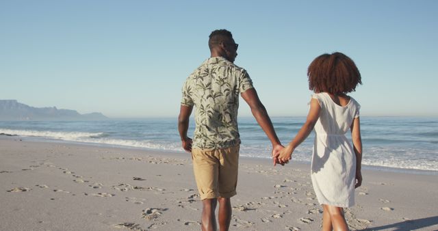 Rear view of happy diverse couple holding hands and walking on sunny beach by the sea. Summer, free time, relaxation, romance and vacations.