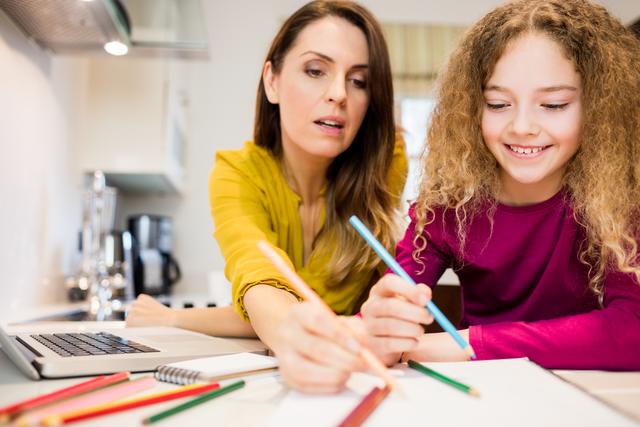 Mother assisting her daughter in drawing at kitchen table. Both are engaged in creative activity with colored pencils and paper. Ideal for concepts related to family bonding, home education, creativity, and parental support. Can be used in articles, blogs, and advertisements focusing on family life, parenting tips, and educational activities at home.
