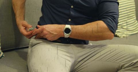 Mature Man Holding Small Gray Remote While Wearing White Smartwatch and Navy Shirt on Sofa