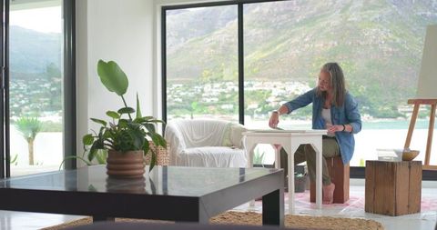 Woman painting by lakeside window in bright minimalist living room with mountain view