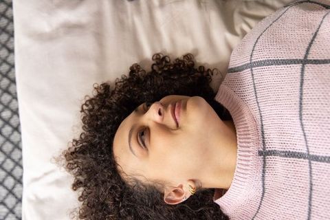 Curly-haired woman lying on bed wearing pink grid sweater and gold hoop earrings