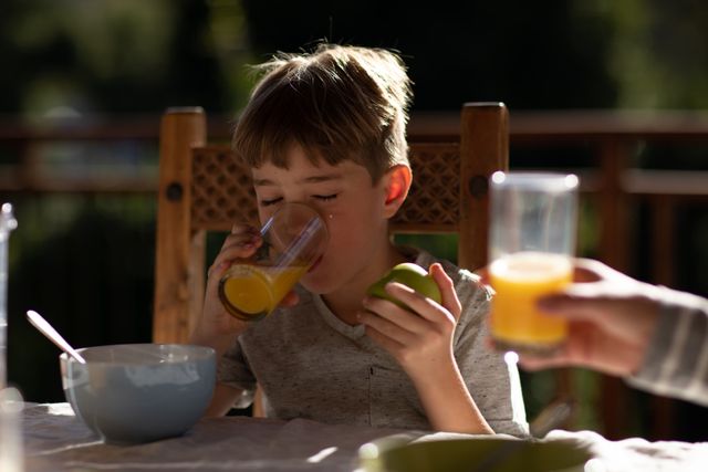 Front view of a Caucasian boy sitting at a table in the garden enjoying breakfast, holding fruit and drinking juice. 