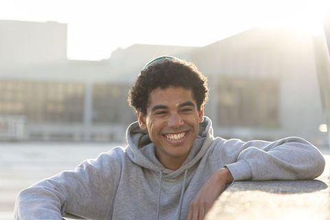 Sunlit teenage male leaning on concrete ledge smiling in gray hoodie with rim light