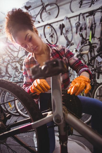 Young female mechanic wearing a plaid shirt and gloves, concentrating while repairing a bicycle in a workshop filled with various bikes and tools. Perfect for illustrating bike maintenance, female professionals in technical fields, and small business operations.