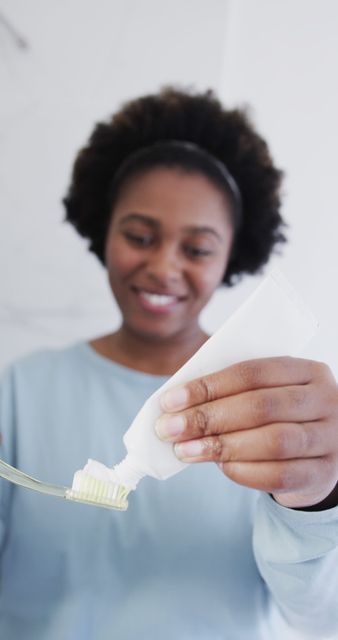 African American woman applying toothpaste on toothbrush, smiling as part of her morning routine. Suitable for content related to oral hygiene, dental care products, healthy lifestyle promotions, and personal care advertisements aimed at diverse audiences.
