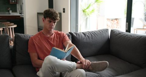 Young man reading on gray sectional sofa in modern bright living room wearing orange tee