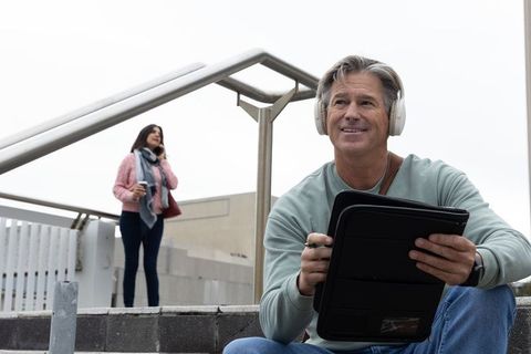 Middle-aged man using tablet with headphones on urban steps while woman talks on phone