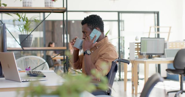 Young African American man multitasking in modern office. Holding two smartphones, using laptop, and drinking coffee. Perfect for illustrating busy work life, productivity tips, and modern business environments.