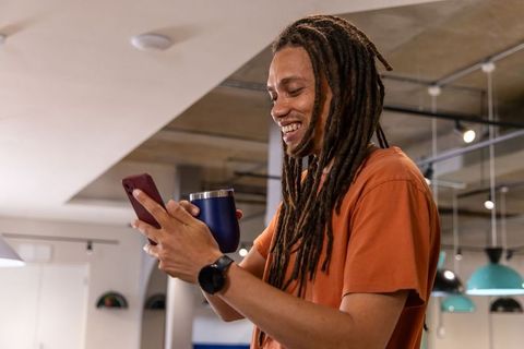 Smiling man with dreadlocks checking phone and holding mug in modern coworking space
