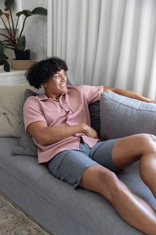 Relaxed african american man reclining on grey sofa in pink shirt, cozy modern living room