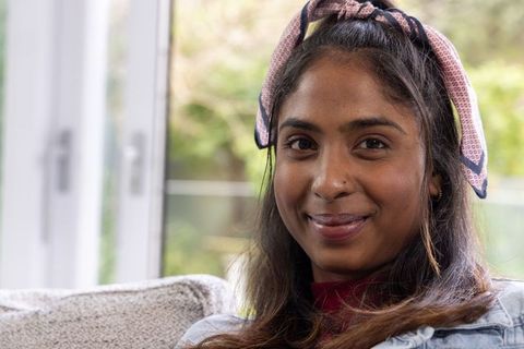 Indian woman in denim jacket wearing patterned headscarf sitting on sofa smiling softly