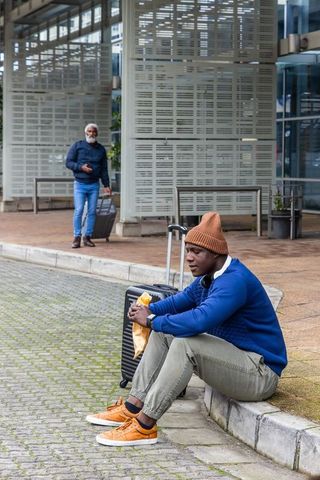 African american man waiting with suitcase eating snack at transit curb urban station