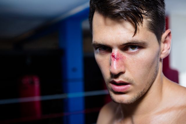 This image captures a close-up of a young male boxer with a bleeding nose, showcasing determination and resilience in a fitness studio. Ideal for use in articles or advertisements related to boxing, combat sports, athletic training, sports injuries, and motivation. It can also be used in fitness blogs, gym promotions, and sportswear advertisements.