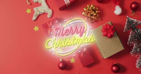 Glowing neon-style merry christmas sign on red flat lay with kraft gift box and baubles