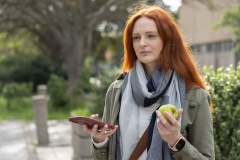 Redhead woman holding phone and apple in urban park wearing scarf and smartwatch