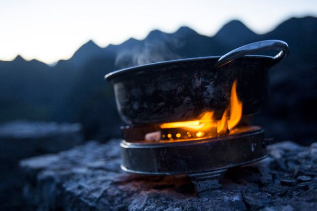 A metal pot is heating on a portable stove in an outdoor setting with mountains silhouetted in the background. Steam rises from the pot, suggesting a hot meal cooking. This image can be used for travel blogs, camping guides, outdoor adventure websites, or advertisements for camping gear.