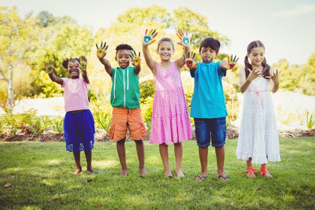 Group of happy children standing in a park with their hands painted in bright colors, smiling and enjoying the outdoors. Ideal for use in advertisements, educational materials, and websites promoting children's activities, creativity, and diversity.