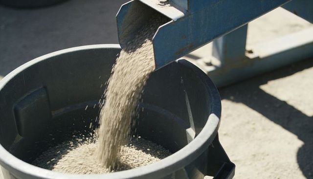 Pouring industrial granules into plastic bin from metal chute under bright sunlight