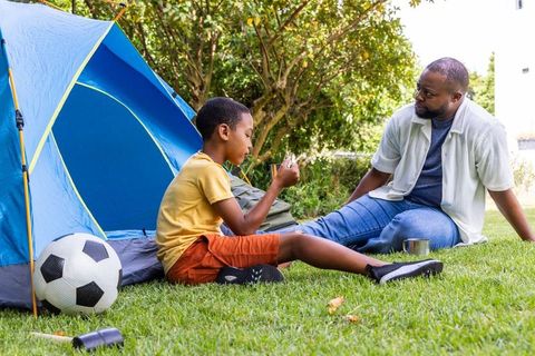 African american father and son camping in backyard examining toy near blue tent