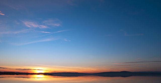 This image shows a serene sunrise with soft golden hues over calm water, reflecting the clear blue sky. Ideal for use in themes surrounding tranquility, nature, morning routines, and peaceful backdrops. It can be used for websites, blogs, and advertisements that emphasize calm and beautiful natural settings.