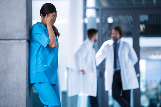 Nurse in blue scrubs leaning against wall, covering face with hand, showing signs of stress and exhaustion. Two doctors conversing in background. Useful for illustrating healthcare worker burnout, mental health challenges in medical field, and stress in healthcare industry.