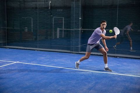 Male player lunging and swinging padel racket on blue court during intense backhand rally