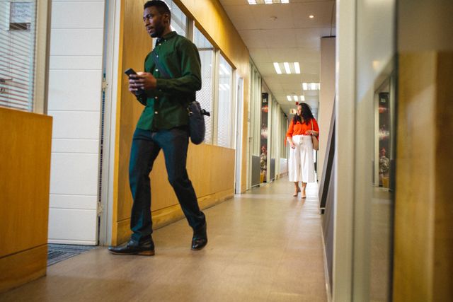 Businessman in smart casual attire walking through an office corridor with a female colleague in the background. Ideal for depicting modern business environments, corporate culture, teamwork, and professional settings. Suitable for use in business websites, corporate presentations, and office-related content.