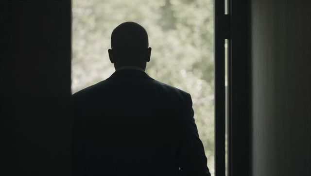 Silhouette of bald man in suit standing at sunlit doorway overlooking lush green foliage