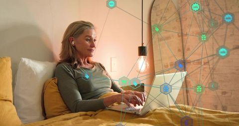 Senior woman reclining in bed with laptop and earbuds, browsing online in cozy warm bedroom