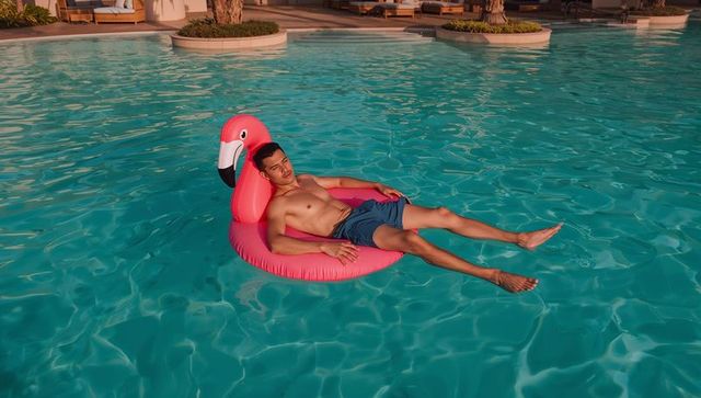 Young man relaxing on pink flamingo pool float at luxury resort pool during golden hour
