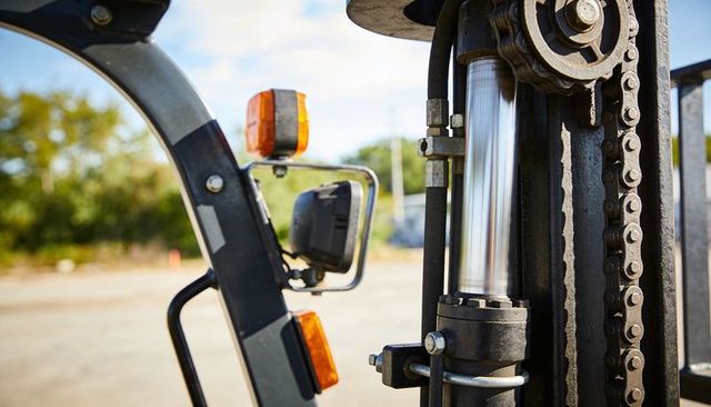 Forklift mast closeup showing chrome hydraulic piston, roller chain, mirror and amber lights
