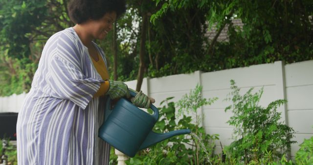 An African American woman is watering plants with a watering can in her home garden. She is dressed in casual clothing and gloves, displaying care for her plants. This could be used in lifestyle blogs, gardening tutorials, articles about hobbies, or promotional material for gardening tools.