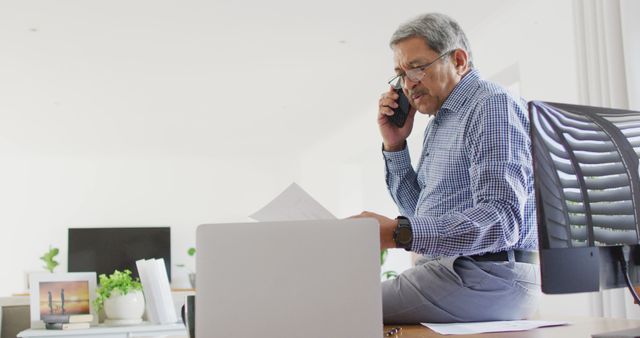 Image of senior biracial man sitting on desk talking on phone and using laptop at home. Business communication, working remotely, inclusivity and senior lifestyle concept.