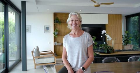Smiling senior woman standing in modern bright open-plan living space wearing smartwatch