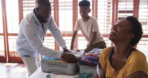 African american family packing suitcases together at home, woman laughing with checklist