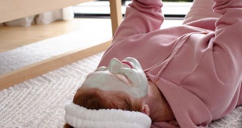 Woman lying on rug wearing facial mask and white headband in cozy pink hoodie relaxing