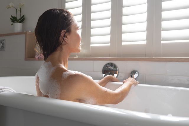 Woman enjoying a relaxing bubble bath at home, looking thoughtful. Ideal for concepts related to self-care, relaxation, wellness, and home lifestyle. Can be used in advertisements for bath products, spa services, or wellness blogs.