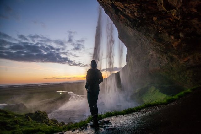Person standing under a cave-like formation, admiring a cascading waterfall at sunset. Lush green moss and wet rocks frame the scene, as dramatic skies add to the natural beauty. Perfect for travel blogs, adventure promotions, or nature-related marketing materials.