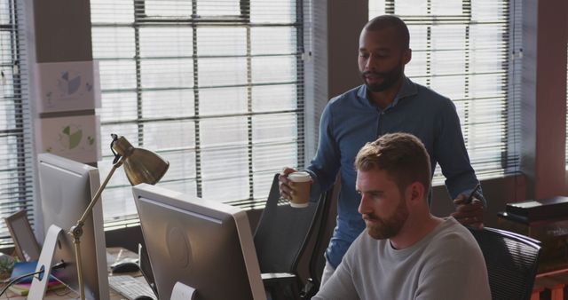 Two colleagues are collaborating on a project in a modern office environment. One person stands holding a coffee cup while communicating with the seated co-worker engaged in work on a computer. This image is useful for illustrating teamwork, professional collaboration, modern workspaces, and discussions in technology or business settings.