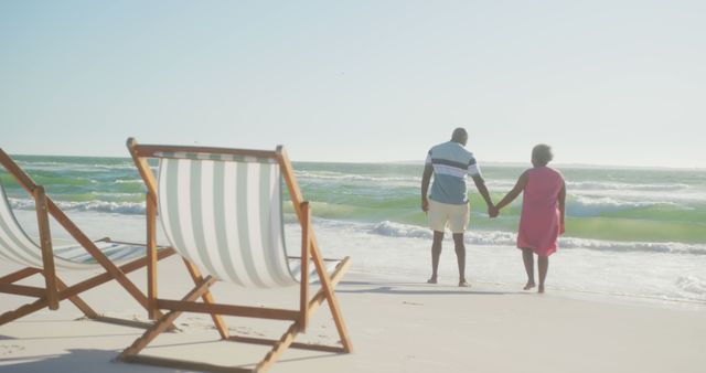 Happy senior african american couple holding hands and walking on sunny beach at sunset, copy space. Retirement, summer, vacations, togetherness and senior lifestyle, unaltered.