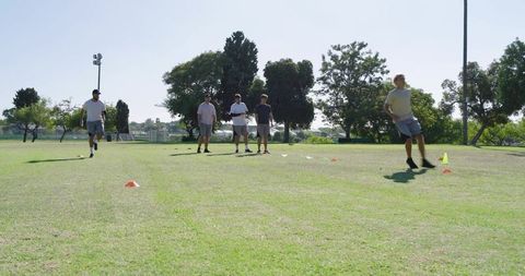 Diverse male athletes running cone agility drill on sunny park field for team training